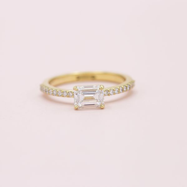 n emerald cut diamond sits in the center of this yellow gold pavé east-west engagement ring.