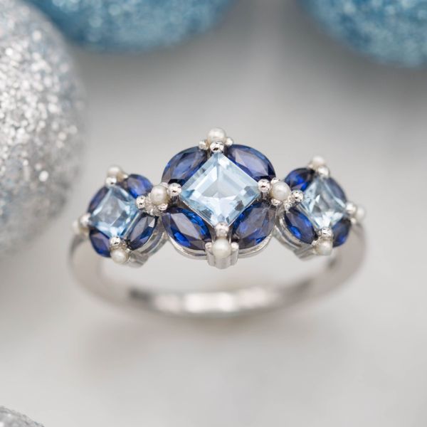 A unique statement ring with three halo-style settings of marquise sapphire and seed pearl around princess cut aquamarines.