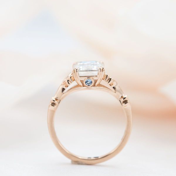 An emerald cut moissanite sits in a double prong rose gold engagement ring setting.