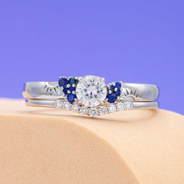Blue sapphire accents of various sizes add beautiful bold pops of color to this moissanite and white gold engagement ring. The deep blue accents reflect in the diamond accents of the matching wedding band.