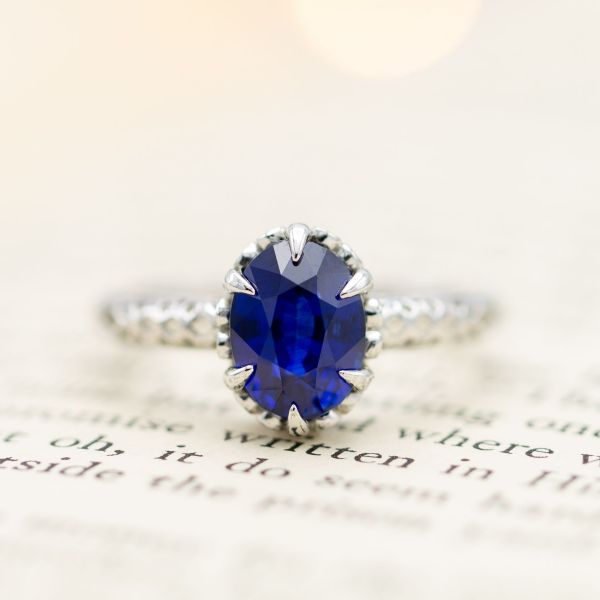 A deep blue, oval cut sapphire engagement ring set with six claw prongs in an intricate filigree setting.