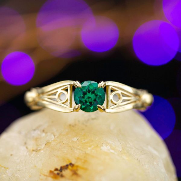 A brilliant round-cut emerald sits between two deathly hallows inspired symbols, between the wings of two familiar gold winged balls.