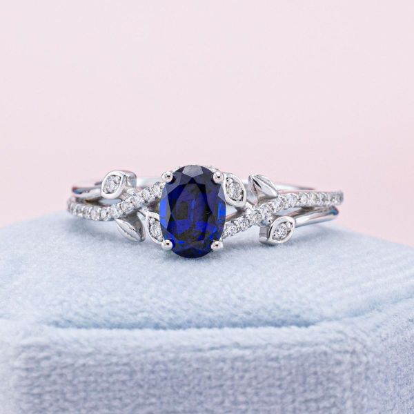 Lab-created diamond accents sparkle around this dark blue, oval, lab-created sapphire. We split the white gold band, embellishing one arm with pavé set diamonds and the other with delicate white gold leaves.