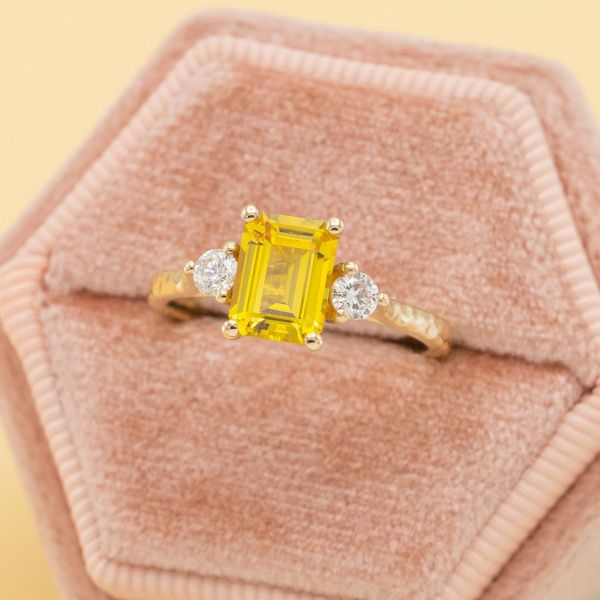 There's sunshine every day with this bright yellow sapphire engagement ring. The emerald cut center stone sits between two round diamond accents in a sunny yellow gold band featuring a hammered metal texture.