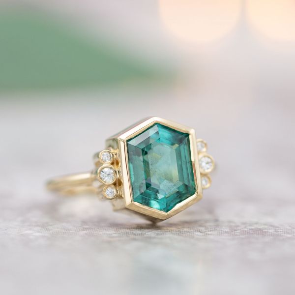 Unique, elongated hexagon cut tourmaline with a blue-green color in a vintage-inspired ring with diamond accents.