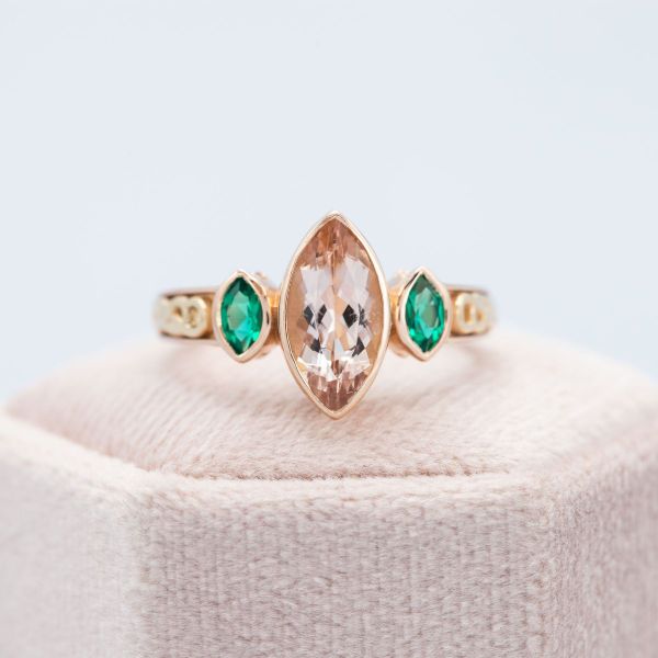 A peach morganite in yellow gold bezel setting with emerald side stones.