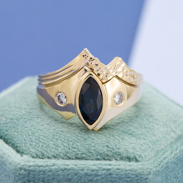 This fantasy-inspired ring boasts a marquise shaped sapphire nestled in a yellow gold bezel with white accent stones and a matching textured wedding band.