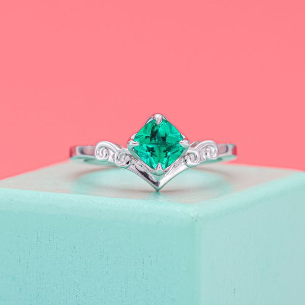 The cushion cut emerald in the center of the white gold tiara shaped engagement ring has a relatively light saturation.