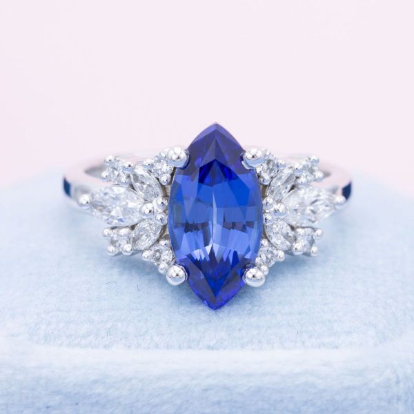 A light blue, lab-created sapphire in a marquise cut shines cooly in the center of this white gold engagement ring. Filigree-like designs made up of round and marquise cut lab diamonds of various sizes flank either side of the center stone.
