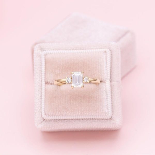 An emerald cut moissanite sits in the center of this yellow gold, three stone engagement ring.