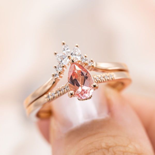 This peachy-pink sapphire and diamond bridal set is both playful and regal at the same time. The beautiful blush of the pear cut sapphire is picked up in the rose gold band. Lab-created diamond accents glisten on either side of the engagement ring and in the tiara wedding band.