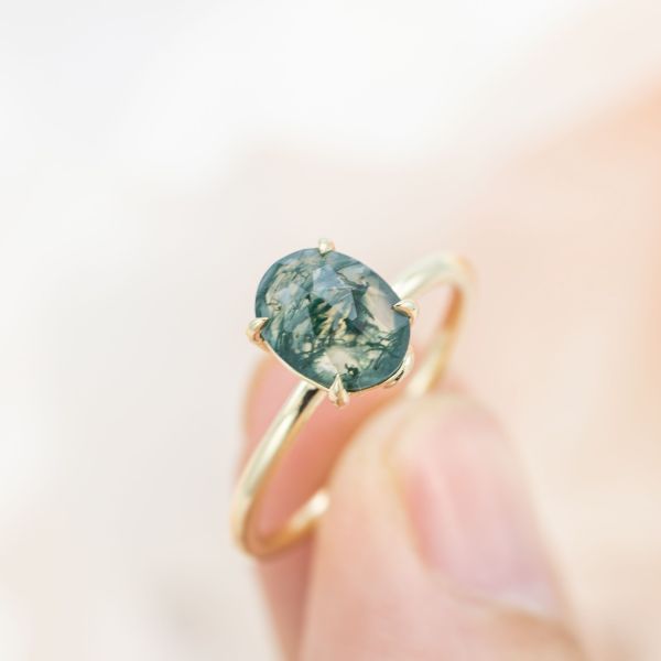 Bright, beautiful solitaire engagement ring with a captivating oval moss agate center stone set with claw prongs on a minimal, delicate, gold band.