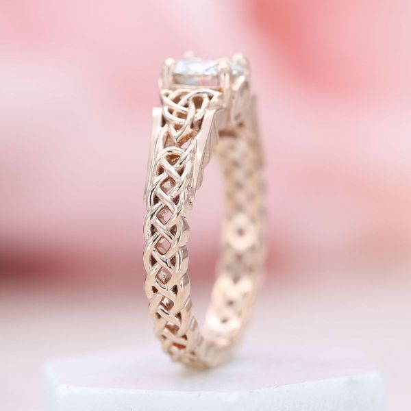 A braided rose gold band weaves into two triquetras supporting a moissanite center stone in this engagement ring.