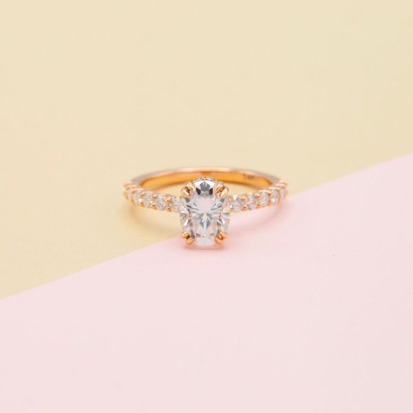 Tapered, claw-like double-prongs contrast with the oval cut moissanite center stone and round moissanite accents adorning this rose gold engagement ring.
