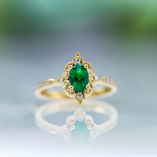 An oval cut emerald sits in the center of this vintage inspired yellow gold engagement ring.