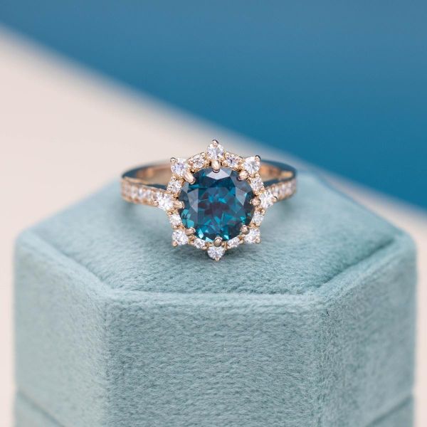 A blue alexandrite sits at the center of a diamond halo in a yellow gold engagement ring.