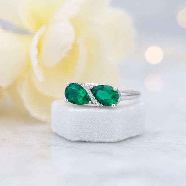 A perfectly elegant pair of pear cut emeralds nestled around a row of sparkling diamond in an east-west arrangement.