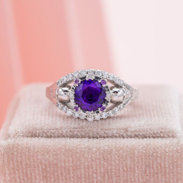 A brilliant round amethyst is flanked by diamond accents in a split shank, white gold engagement ring featuring hidden skull accents.