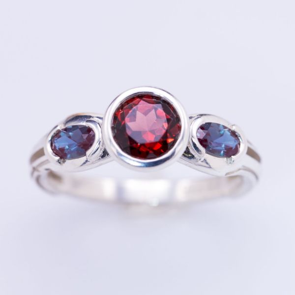 A deep red garnet center stone draws out the purple shades of the bezel set oval alexandrite side stones in this modern ring.