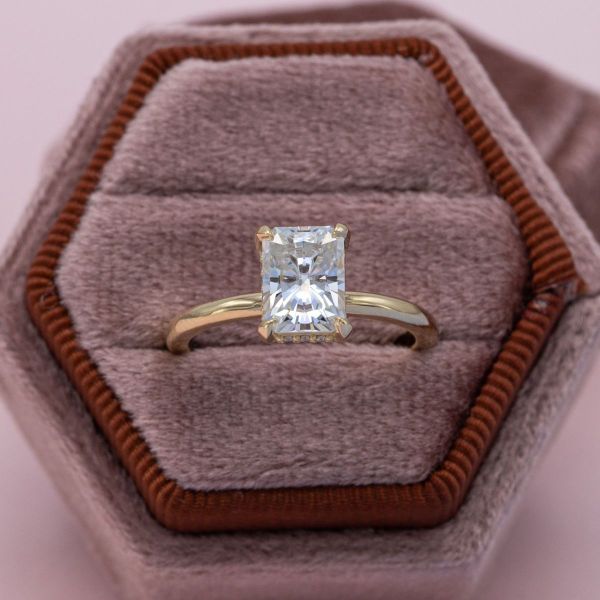 A hidden halo wraps around the pavilion of the emerald cut moissanite center stone, set on a simple yellow gold band.