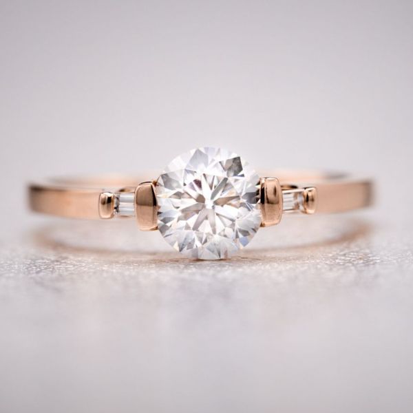 1.01克拉的RBC钻石镶嵌在现代玫瑰金条上，镶有长棍形侧边宝石，戒指内隐藏着两颗宝石(一颗祖母绿和一颗蓝宝石)。